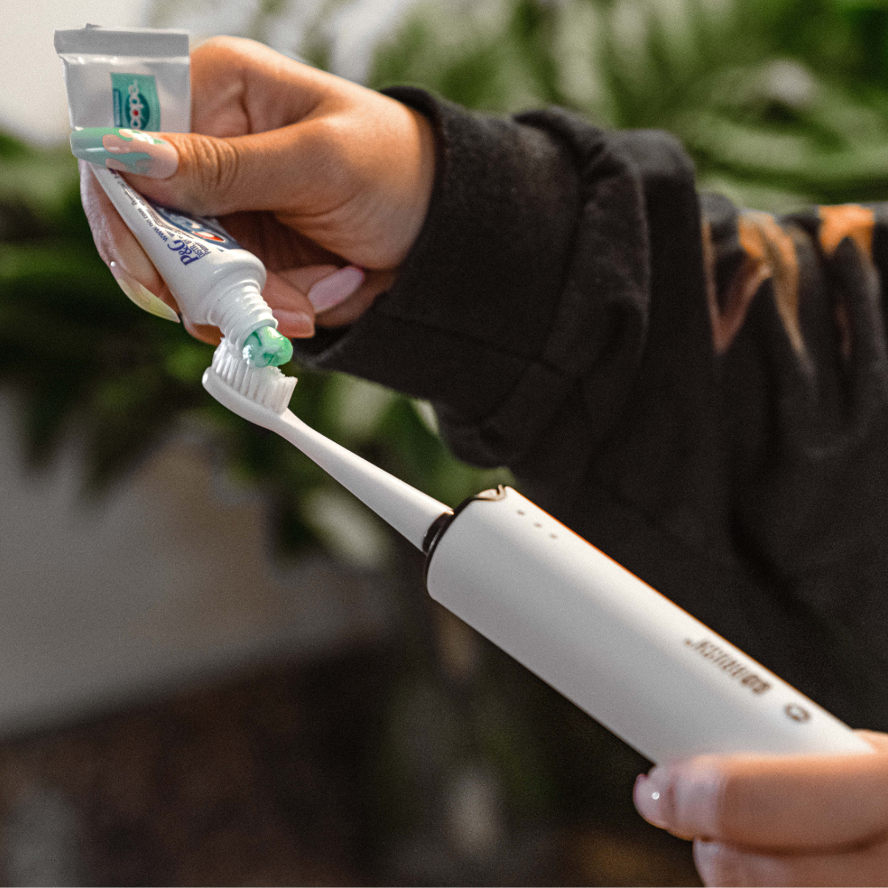 Person applying toothpaste to a white electric toothbrush with a blurred background