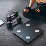 Person sitting on a digital weight scale with dumbbells and a phone nearby on a dark floor.