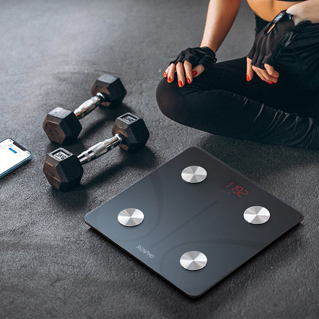 Person sitting on a digital weight scale with dumbbells and a phone nearby on a dark floor.