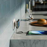 A smartphone lies on a marble kitchen countertop, connected to a wall charger plugged into an outlet. A tablet rests on a wooden cutting board with a knife nearby, and a sink and tiled wall are blurred in the background.