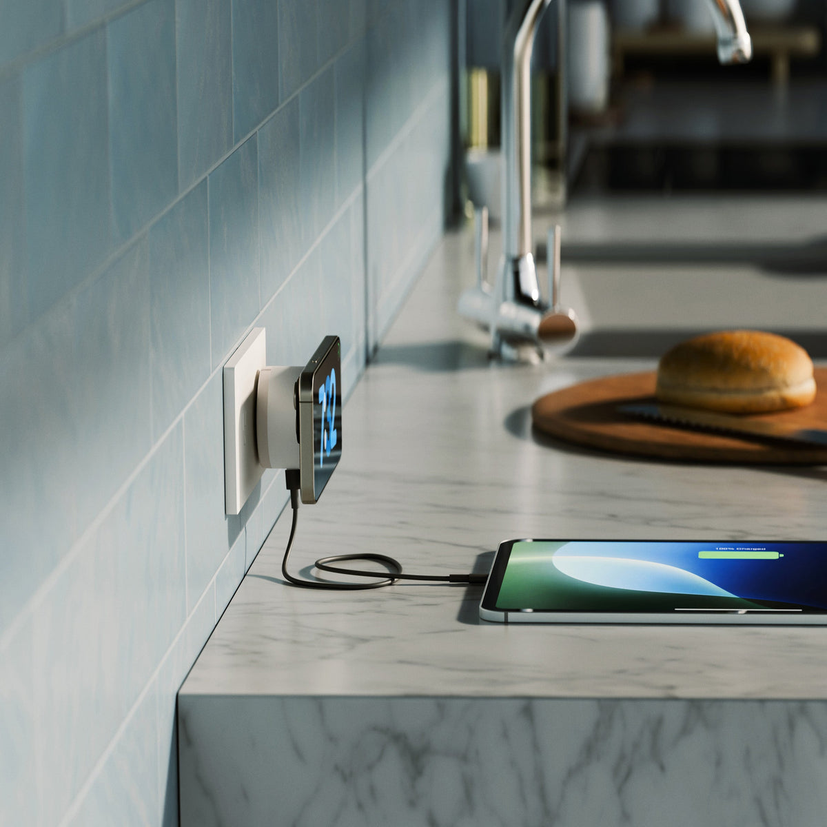 A smartphone lies on a marble kitchen countertop, connected to a wall charger plugged into an outlet. A tablet rests on a wooden cutting board with a knife nearby, and a sink and tiled wall are blurred in the background.