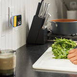 A small digital kitchen timer hangs on a white subway tile backsplash next to a black knife block and a person chopping green herbs on a white cutting board.