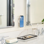 A smartphone mounted on a wall in a bright bathroom, displaying a blue digital clock or calendar app. The bathroom features a white sink, chrome faucet, mirrors, a small tray with eyeglasses, toothbrushes, soap, and a vase with green stems.