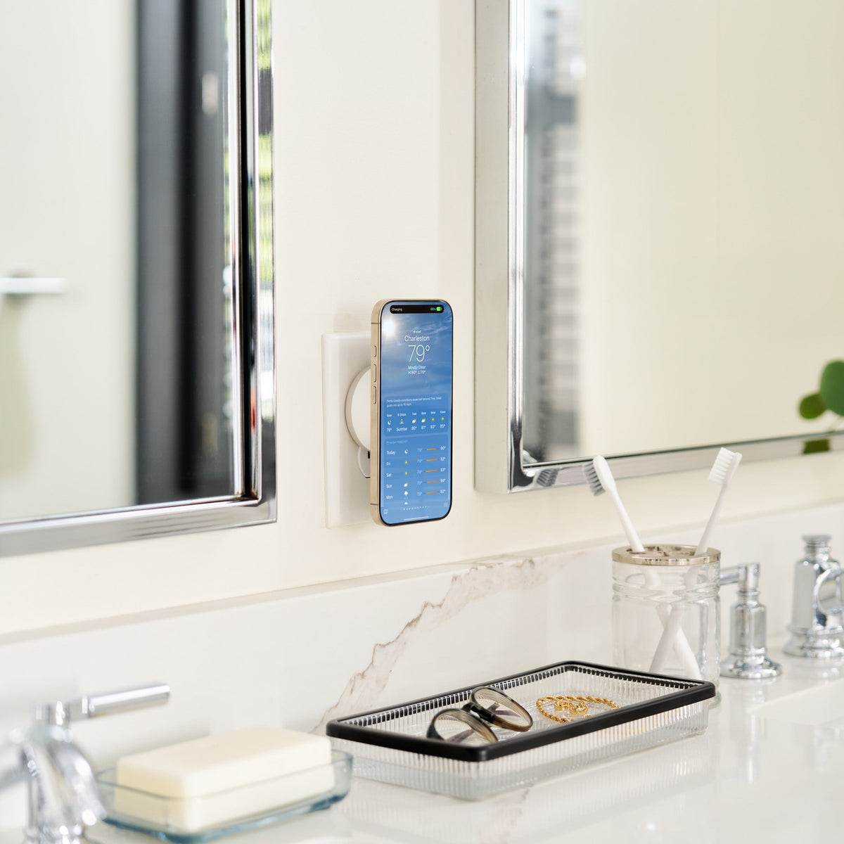 A smartphone mounted on a wall in a bright bathroom, displaying a blue digital clock or calendar app. The bathroom features a white sink, chrome faucet, mirrors, a small tray with eyeglasses, toothbrushes, soap, and a vase with green stems.